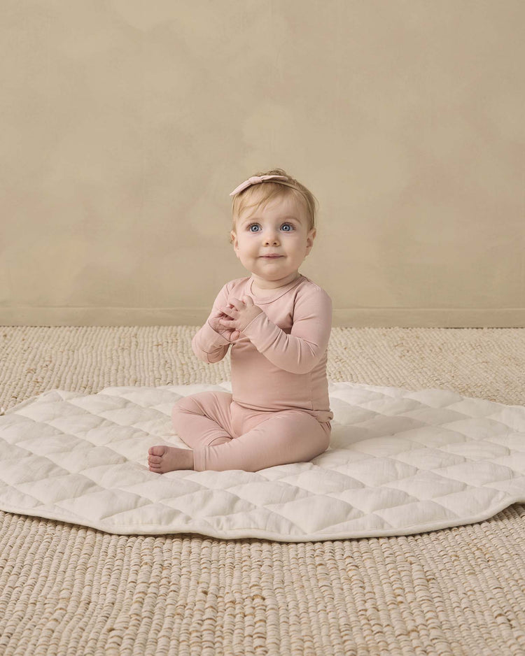 A baby wearing the quincy mae bamboo pajama set in dusty rose, paired with a matching headband, sits on a quilted cream mat with hands clasped, looking up. The background features a beige textured rug.