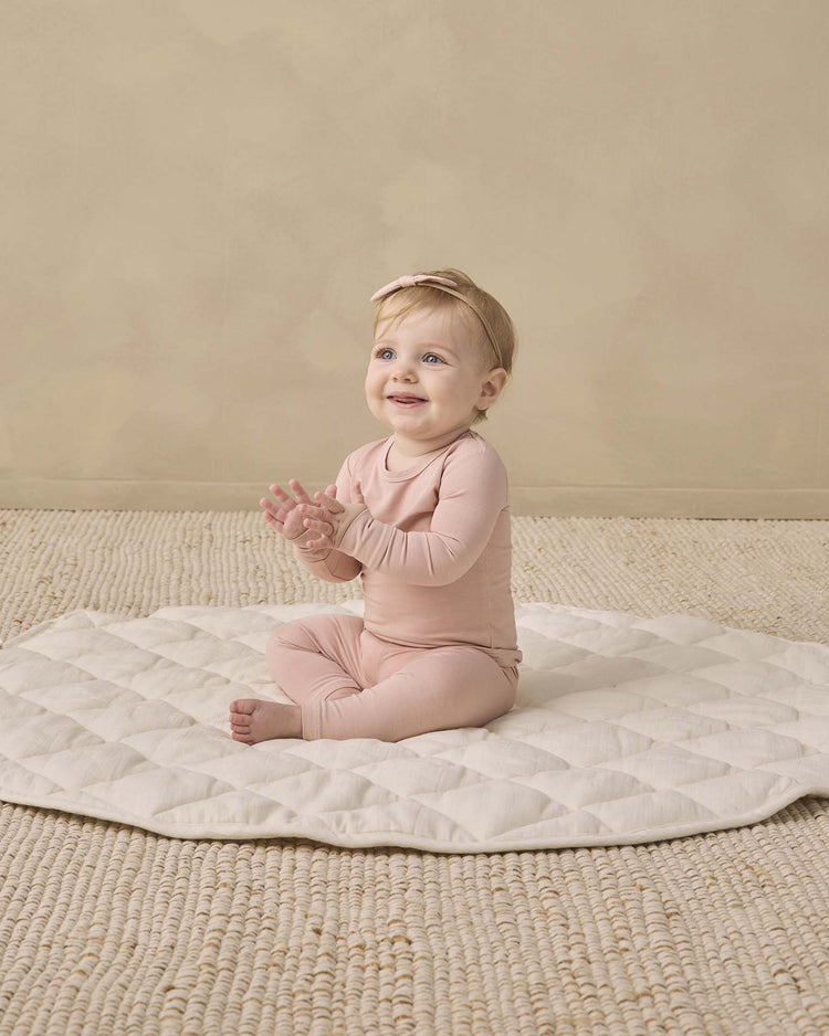 A baby wearing the quincy mae bamboo pajama set in dusty rose and a matching headband sits on a quilted mat, clapping hands and smiling, on a beige textured floor with a plain beige background.