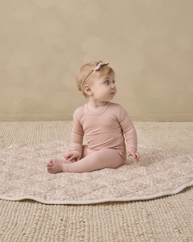 A baby wearing the quincy mae bamboo pajama set in dusty rose with a matching headband sits on a patterned mat, looking to the side, on a textured beige floor against a light tan wall.