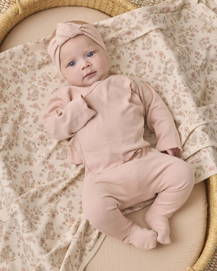 A baby wearing the quincy mae wrap top + footed pant set in dusty rose, complete with a matching headwrap, lies on a beige floral-patterned blanket in a woven basket.