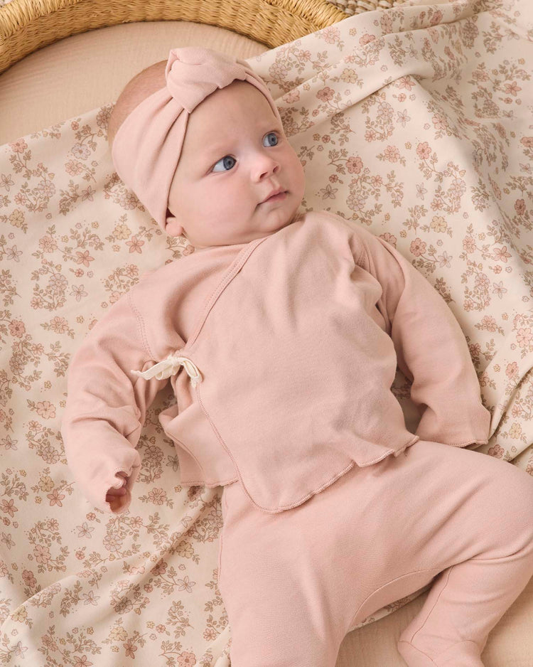 A baby wearing the quincy mae wrap top + footed pant set in dusty rose, along with a matching pink headband, lies on a cream-colored blanket with a floral pattern.