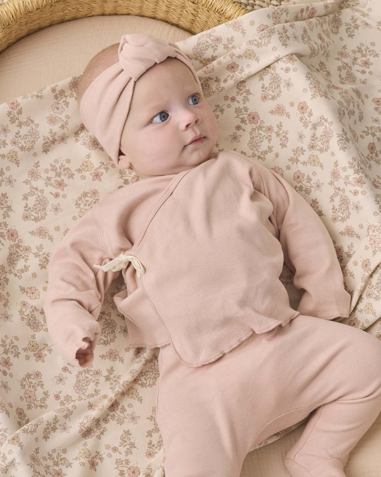 A baby wearing the quincy mae wrap top + footed pant set in dusty rose, along with a matching pink headband, lies on a cream-colored blanket with a floral pattern.