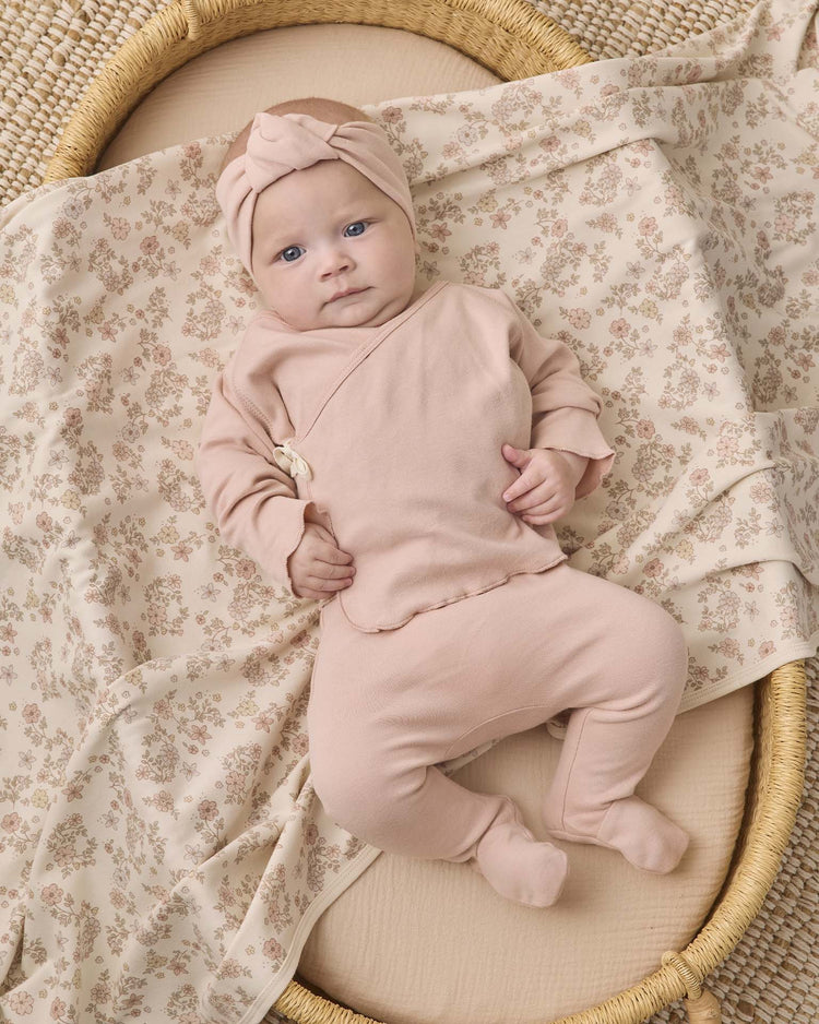 A baby is lying in a woven basket, wearing a pink headband and the Quincy Mae wrap top + footed pant set in dusty rose, on a soft floral-patterned blanket.