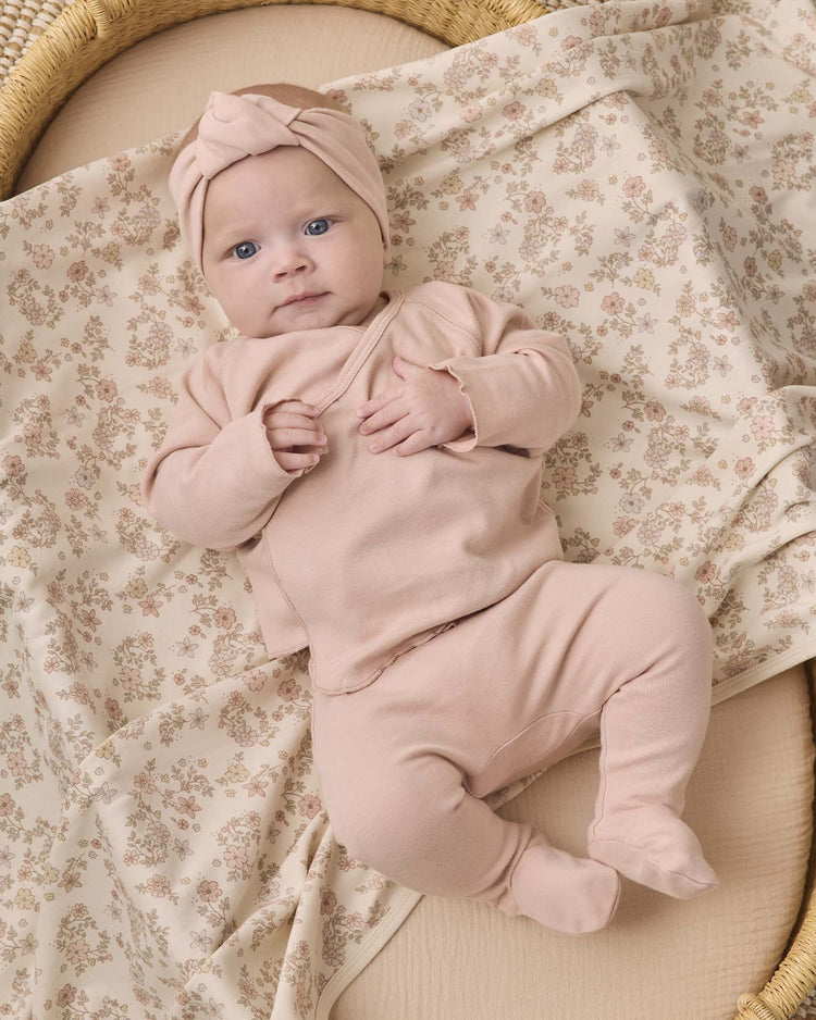 A baby in the quincy mae wrap top + footed pant set in dusty rose and a matching headwrap lies on a cream floral blanket, gazing up with blue eyes and holding their shirt. The organic cotton baby clothes from quincy mae add to the soft, cozy feel of the scene.