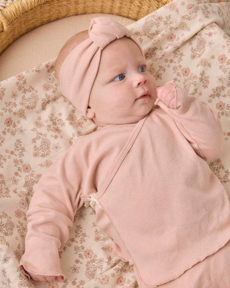 A baby lies on a floral-patterned blanket, wearing the quincy mae wrap top + footed pant set in dusty rose with a matching headband, gazing off to the side with a curious expression.