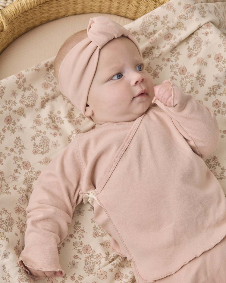 A baby lies on a floral-patterned blanket, wearing the quincy mae wrap top + footed pant set in dusty rose with a matching headband, gazing off to the side with a curious expression.