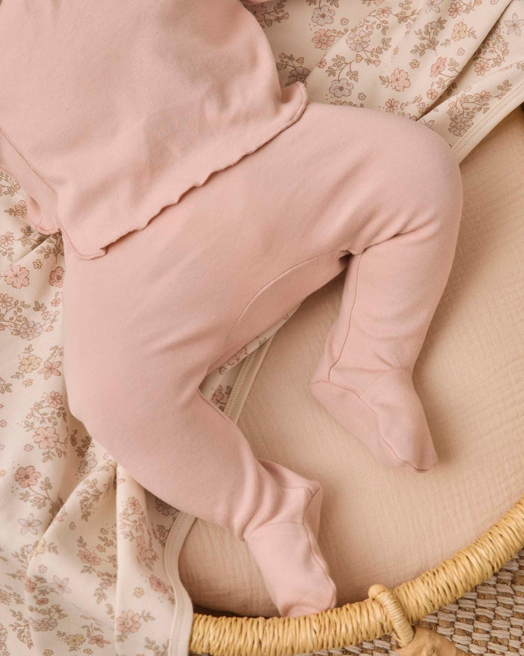A baby wearing the quincy mae wrap top + footed pant set in dusty rose lies on a cream floral-patterned blanket in a woven basket. Only the baby's lower body and part of the adorable newborn wrap top are visible.