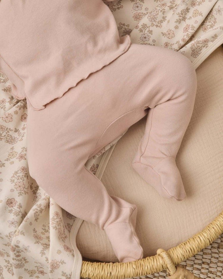 A baby wearing the quincy mae wrap top + footed pant set in dusty rose lies on a cream floral-patterned blanket in a woven basket. Only the baby's lower body and part of the adorable newborn wrap top are visible.