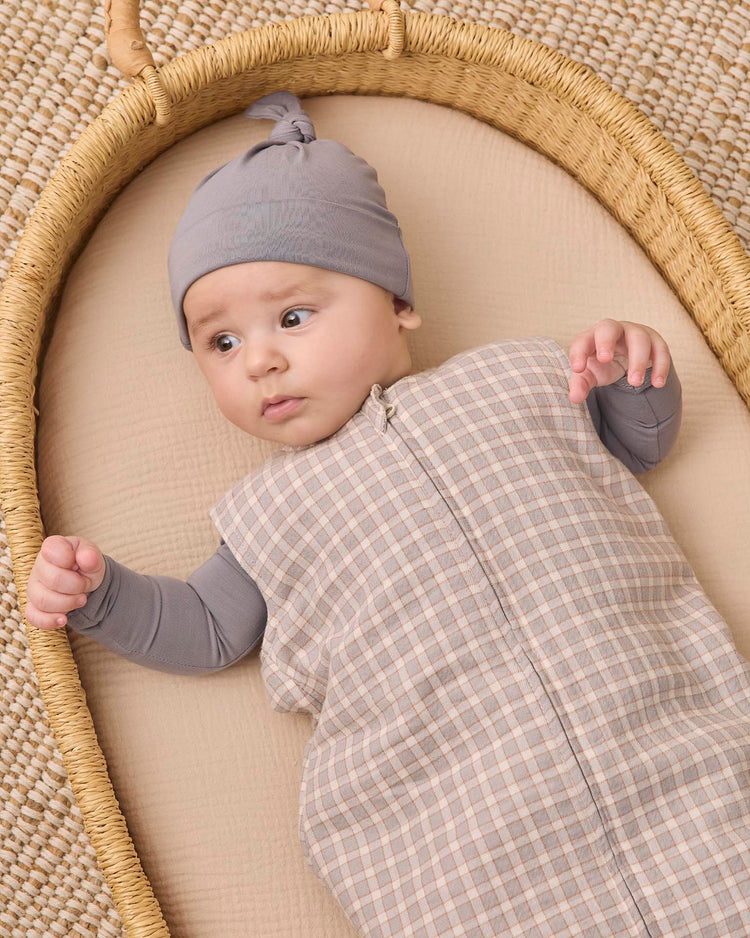 A baby wearing a gray hat and the quincy mae sleep bag in blue gingham, made from organic cotton, lies on a beige cushion in a woven basket, gazing to the side with one arm outstretched.