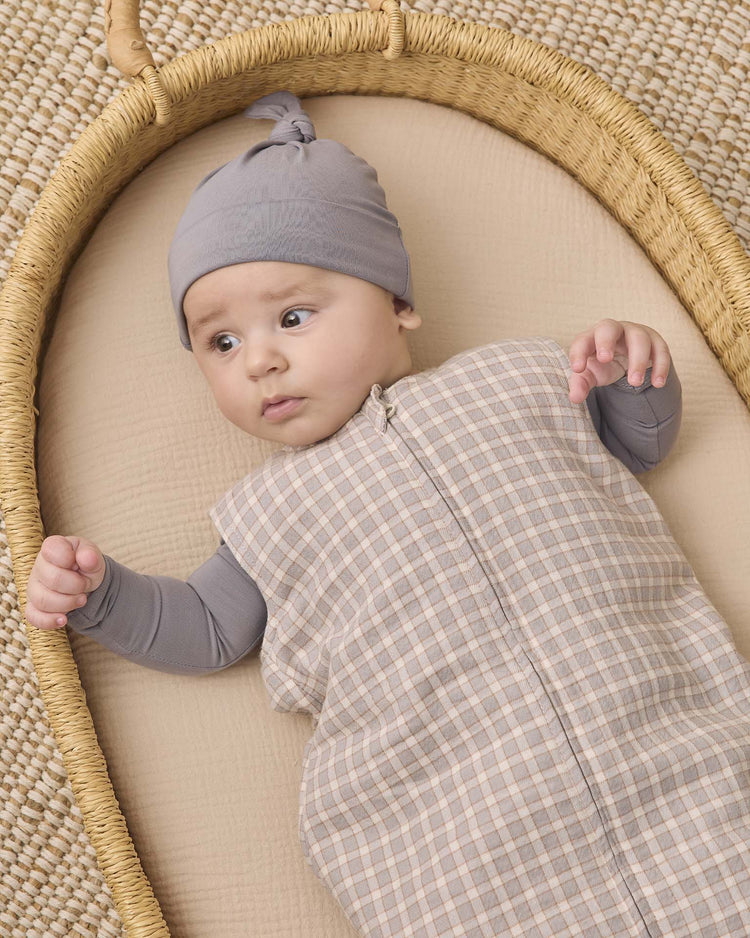 A baby wears the quincy mae bamboo knotted baby hat in slate while lying in a woven basket on a beige mat, looking off to the side with one arm raised.