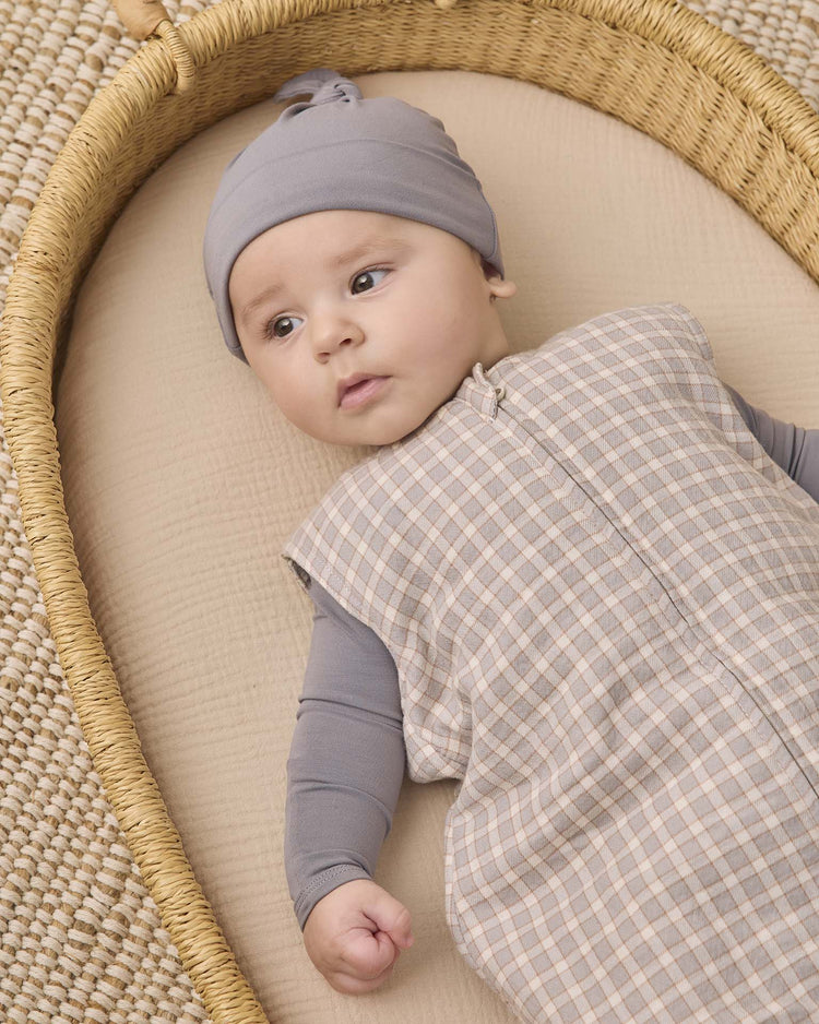 A baby wearing a gray hat and the quincy mae sleep bag in blue gingham lies on a beige mattress in a woven basket, gazing calmly off to the side.