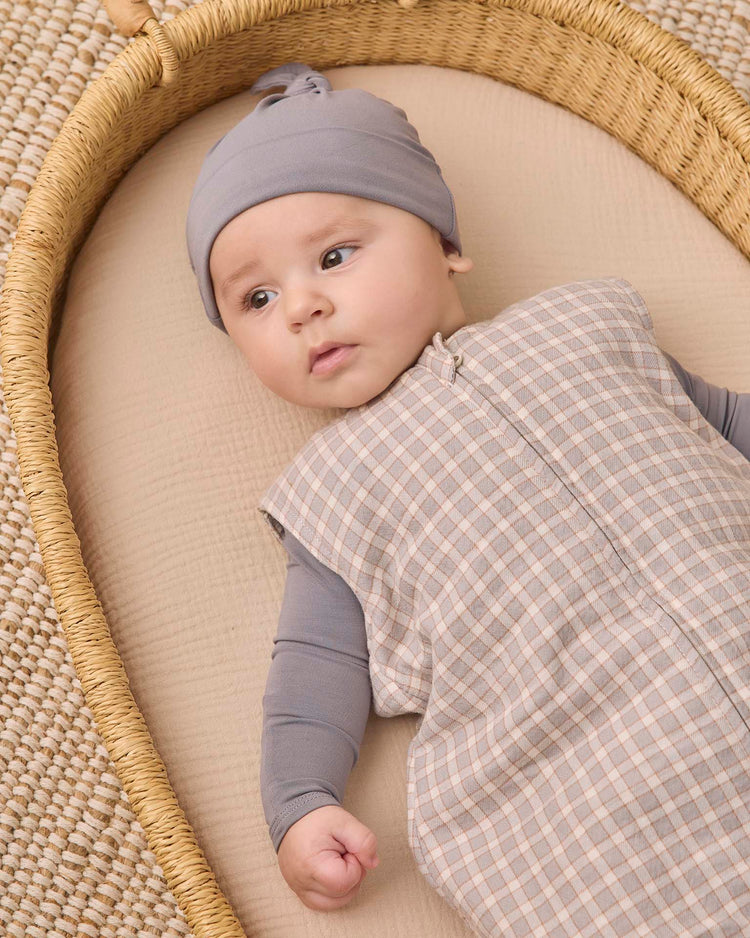 A baby wearing a gray hat and the quincy mae sleep bag in blue gingham lies on a beige mattress in a woven basket, gazing calmly off to the side.