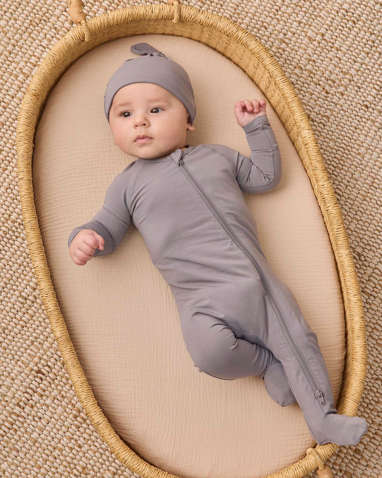 A baby in a quincy mae bamboo zip footie in slate and matching hat lies on a woven oval mat, resting on a textured beige surface.