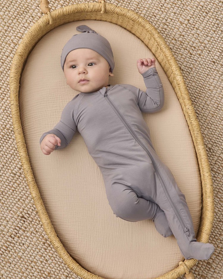 A baby in a quincy mae bamboo zip footie in slate and matching hat lies on a woven oval mat, resting on a textured beige surface.