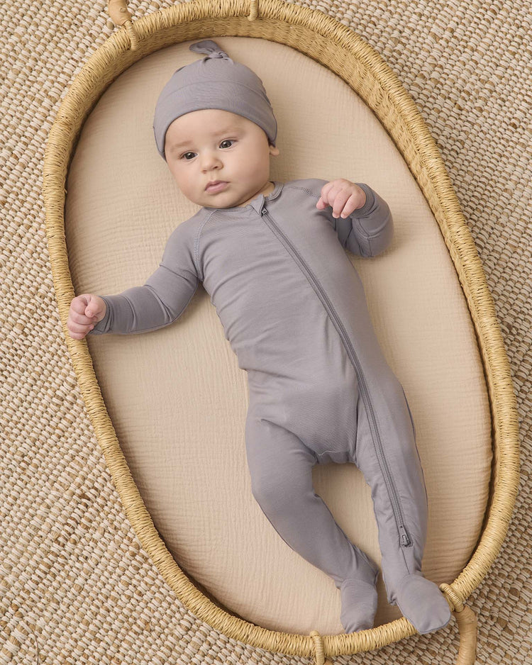 A baby wearing the quincy mae bamboo zip footie in slate and a matching hat lies in an oval woven basket on a textured beige surface, looking up with one arm raised.