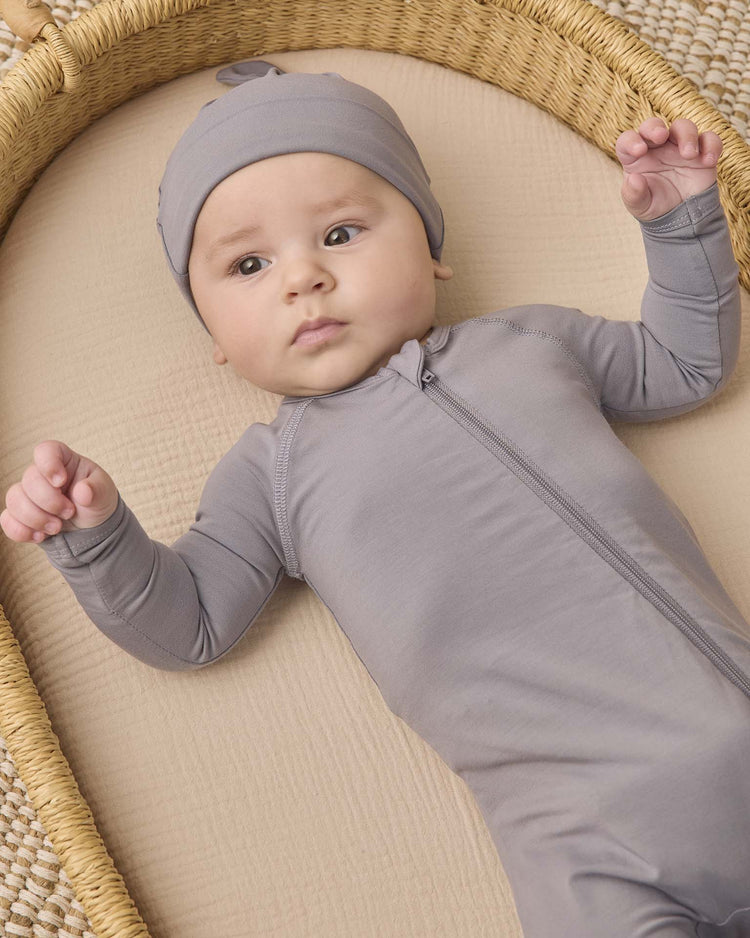 A baby wearing the quincy mae bamboo zip footie in slate with a matching hat lies on a beige mattress in a woven basket, looking up with arms slightly raised. This cozy piece of baby sleepwear features a practical double-zip design for easy changes.