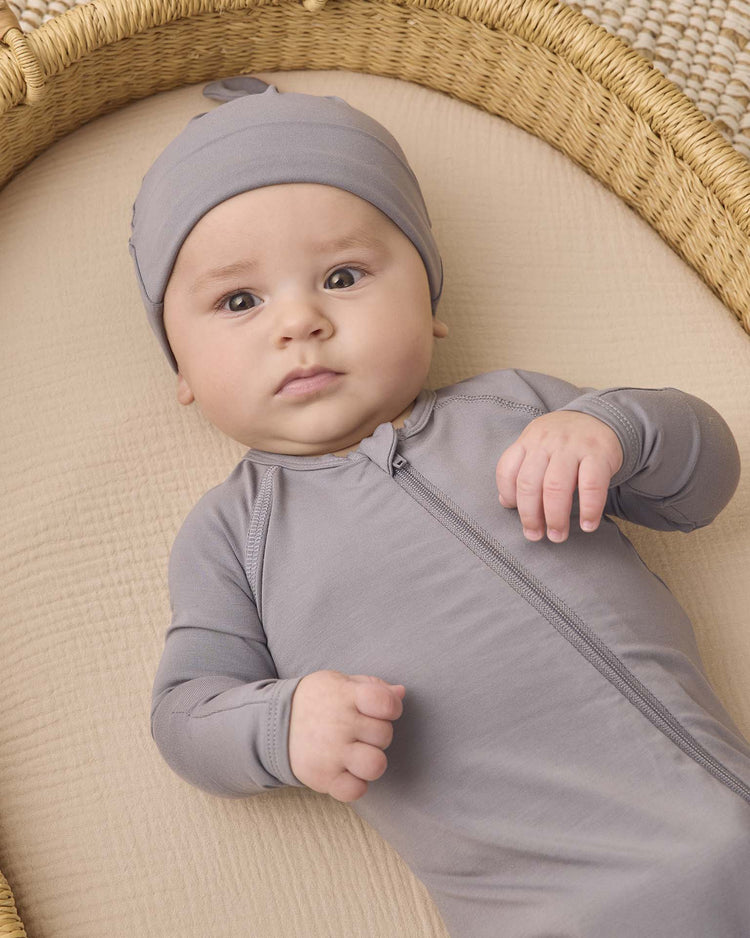 A baby wearing a soft, gray zip-up onesie and the quincy mae bamboo knotted baby hat in slate lies on a beige surface inside a woven basket, looking up with a neutral expression.