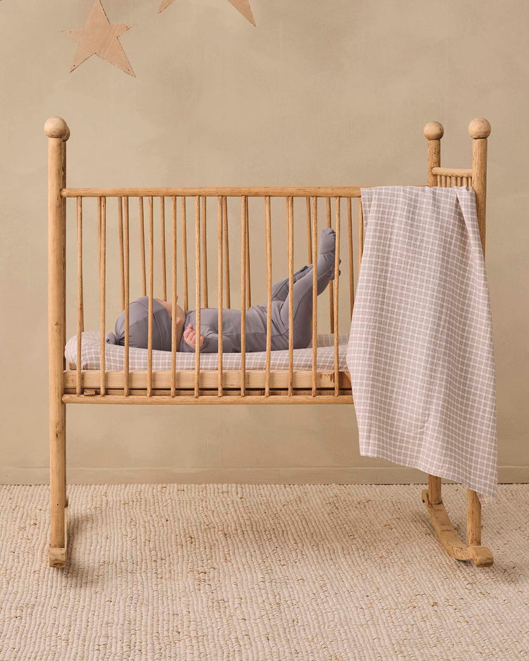 A baby wearing the quincy mae bamboo zip footie in slate lies on their back in a wooden crib with a checkered blanket draped over the side. The cozy double-zip design makes changes easy, while the room features a neutral color scheme and two star decorations on the wall.
