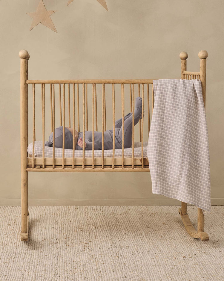 A baby wearing the quincy mae bamboo zip footie in slate lies on their back in a wooden crib with a checkered blanket draped over the side. The cozy double-zip design makes changes easy, while the room features a neutral color scheme and two star decorations on the wall.