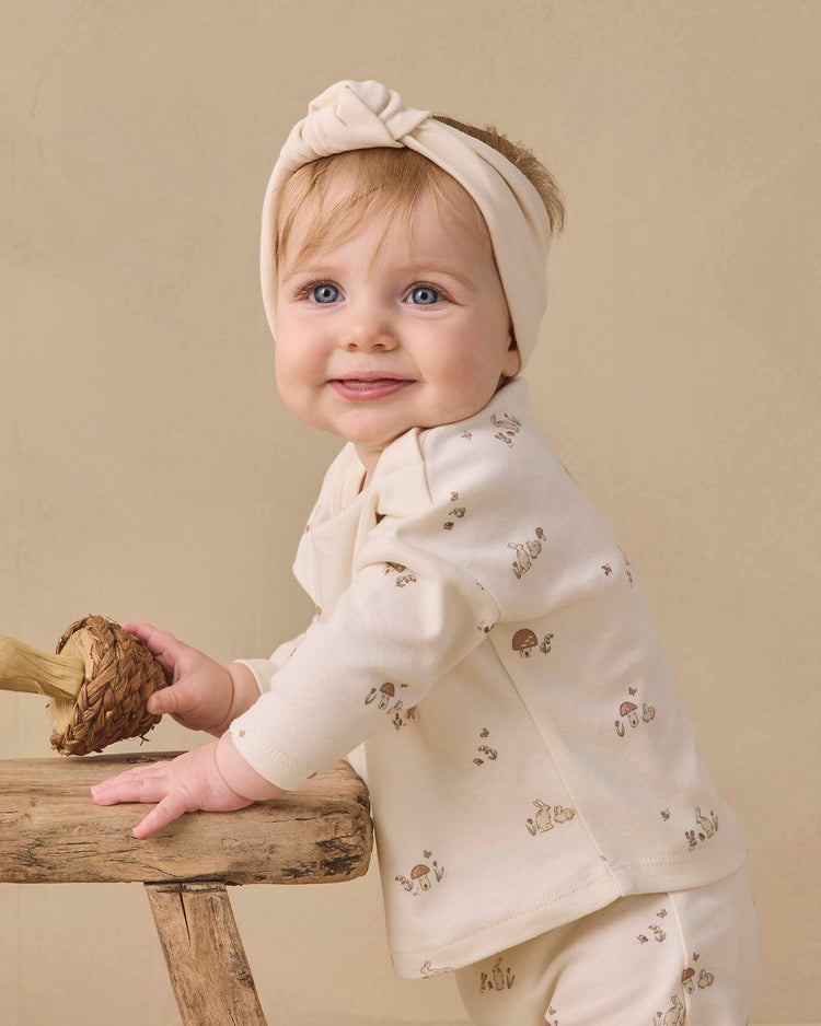 A smiling baby wearing a cream-colored outfit and the quincy mae knotted headband in natural stands, holding a small woven object on a rustic wooden stool against a beige background.