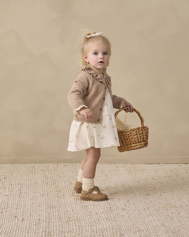 A young girl wears the quincy mae tiered jersey dress in bunnies, paired with a brown cardigan and standing on a textured rug against a beige background. She has blonde hair tied with a bow and finishes the look with socks and brown shoes, holding a wicker basket.