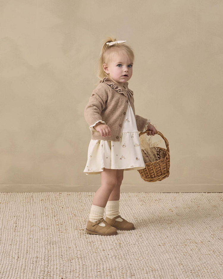 A young girl with blonde hair in a ponytail wears the quincy mae tiered jersey dress in bunnies, paired with a tan cardigan, organic cotton socks and shoes, holding a wicker basket, standing on a textured rug against a beige wall.