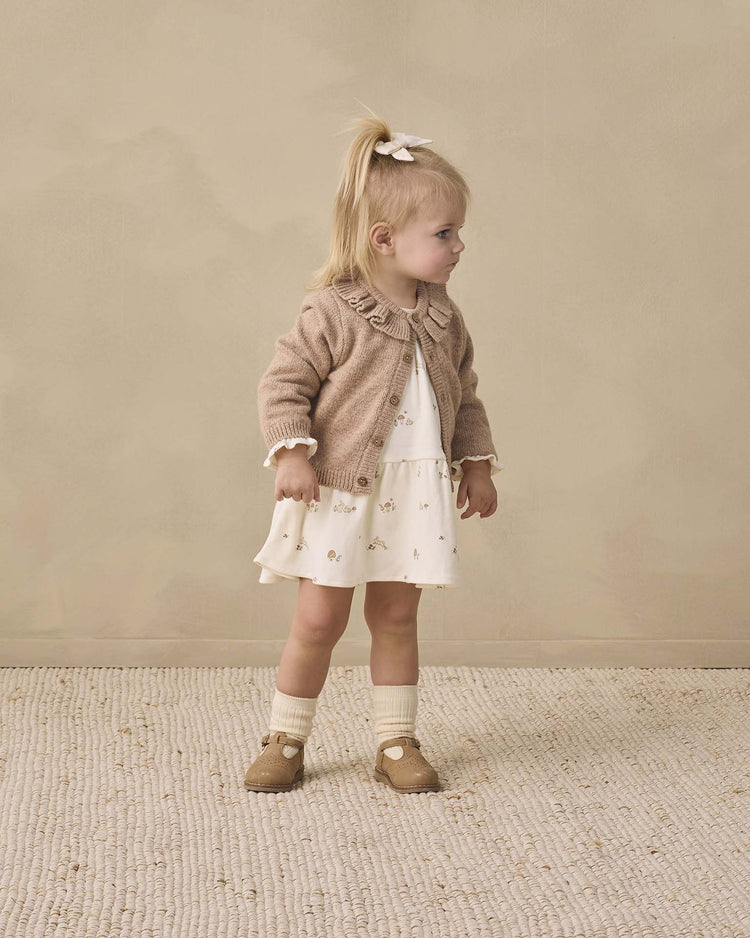 A young girl with blonde hair in a ponytail wears the Quincy Mae tiered jersey dress in bunnies, paired with a beige cardigan, white socks, and tan shoes. She stands on a textured rug, looking to the side against a neutral background.