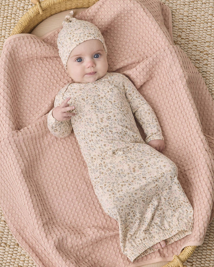 A baby wearing the quincy mae bamboo baby gown + hat set in bloom, featuring fold-over hand mitts and a matching floral hat, lies on a textured, light pink blanket in a woven basket, gazing up with bright blue eyes and a slight smile.