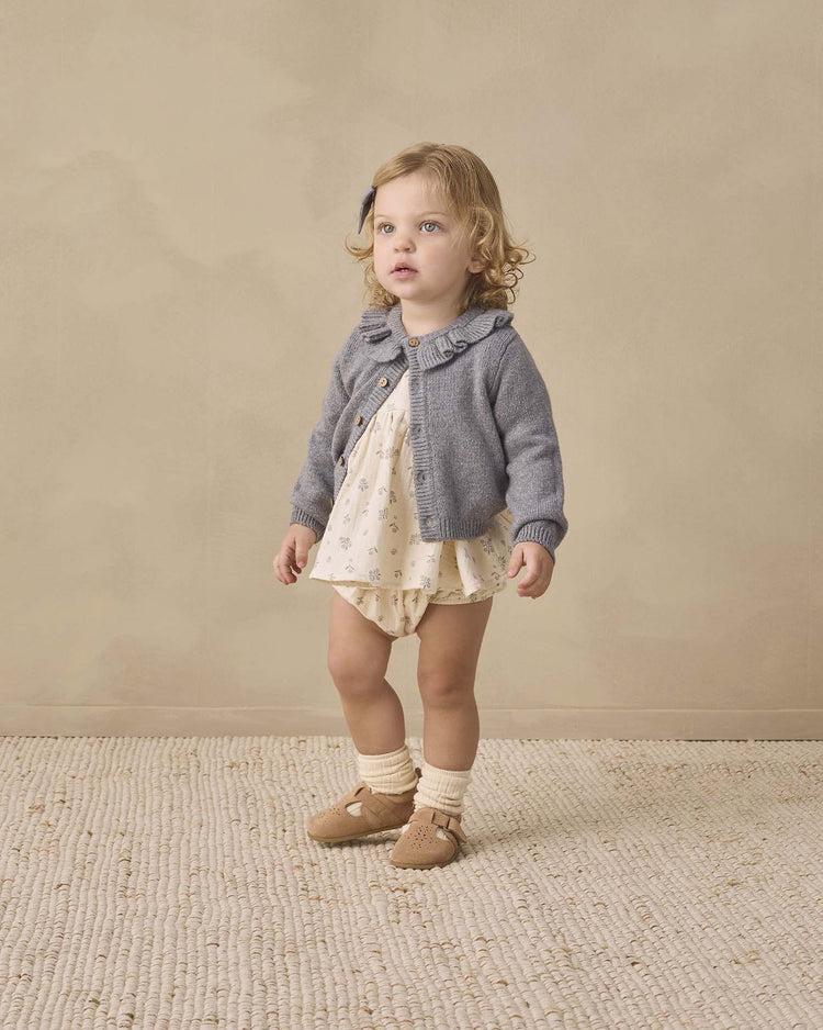 A young child with curly blonde hair stands on a textured rug, wearing the quincy mae francy set in winter berry—an organic cotton baby ensemble—paired with a light romper, gray cardigan, and beige shoes with socks, looking slightly upward against a plain beige background.