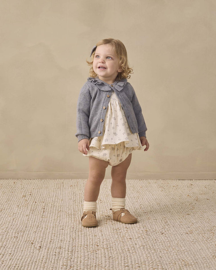 A toddler stands smiling, dressed in the quincy mae francy set in winter berry—an organic cotton baby outfit featuring a blue cardigan layered over a cream empire waist top and matching bloomers, paired with socks and tan shoes. The background is neutral, and the floor is textured.