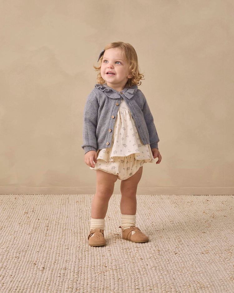 A toddler stands smiling, dressed in the quincy mae francy set in winter berry—an organic cotton baby outfit featuring a blue cardigan layered over a cream empire waist top and matching bloomers, paired with socks and tan shoes. The background is neutral, and the floor is textured.