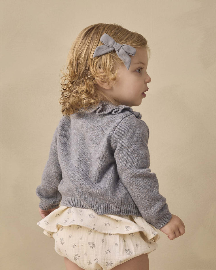 A young child with blonde curly hair, dressed in the Quincy Mae Francy Set in Winter Berry—a cozy, organic cotton ensemble featuring an empire waist top and cream ruffled floral baby bloomers—pairs it stylishly with a matching gray hair bow, standing against a plain beige background.
