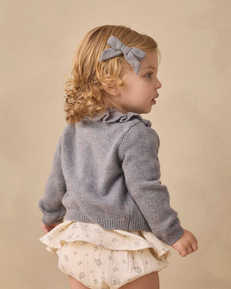 A young child with blonde curly hair, dressed in the Quincy Mae Francy Set in Winter Berry—a cozy, organic cotton ensemble featuring an empire waist top and cream ruffled floral baby bloomers—pairs it stylishly with a matching gray hair bow, standing against a plain beige background.