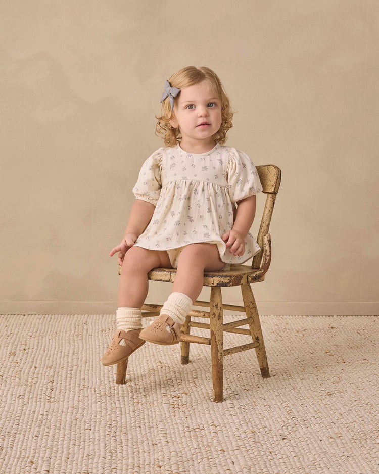 A young toddler with curly blonde hair, wearing the quincy mae francy set in winter berry, sits on a small wooden chair in a neutral-colored room, looking toward the camera.