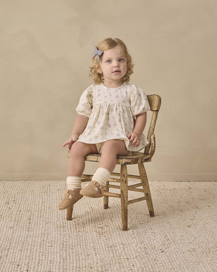 A young toddler with curly blonde hair, wearing the quincy mae francy set in winter berry, sits on a small wooden chair in a neutral-colored room, looking toward the camera.