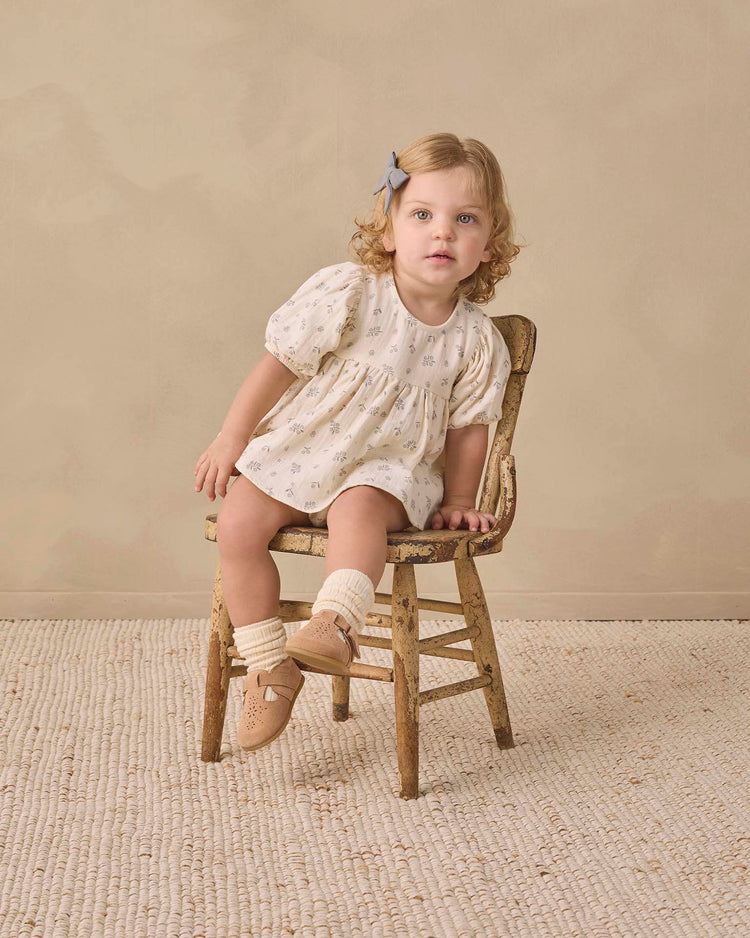 A toddler with curly blonde hair and a blue bow sits on an old wooden chair, wearing the quincy mae francy set in winter berry, paired with beige socks and tan shoes, on a textured rug against a beige background.