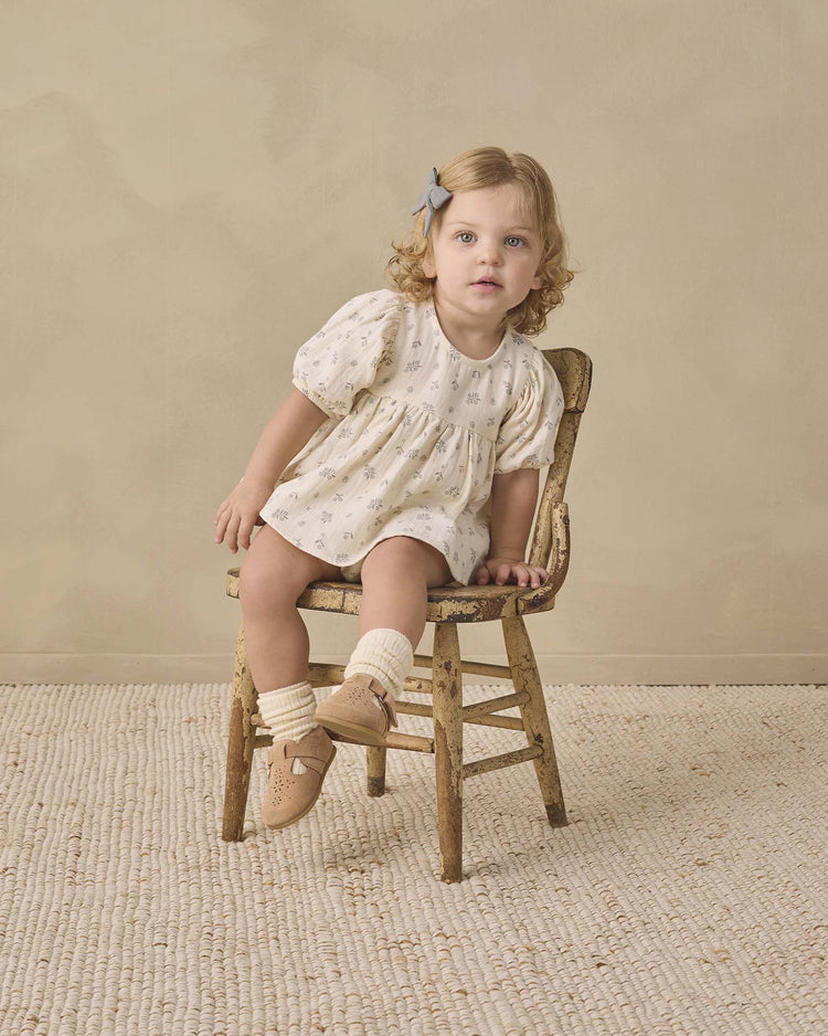 A toddler with curly blonde hair and a blue bow sits on an old wooden chair, wearing the quincy mae francy set in winter berry, paired with beige socks and tan shoes, on a textured rug against a beige background.