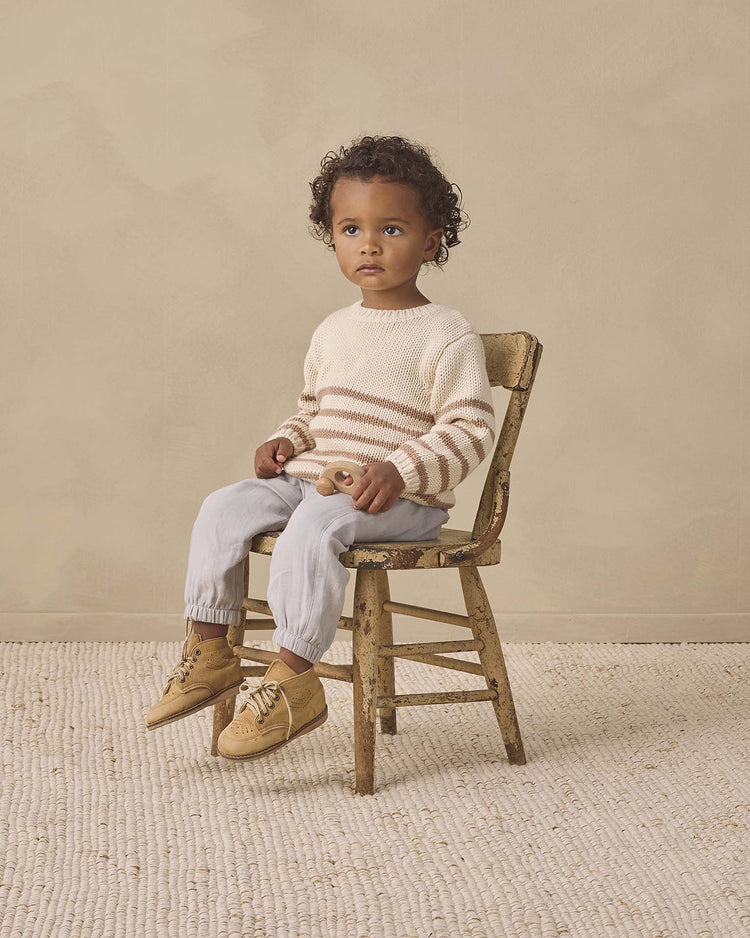 A young child with curly hair sits on a wooden chair, wearing the quincy mae rex sweater in almond stripe crafted from cozy organic cotton, paired with light blue pants and tan shoes. The background is beige with a textured rug on the floor.