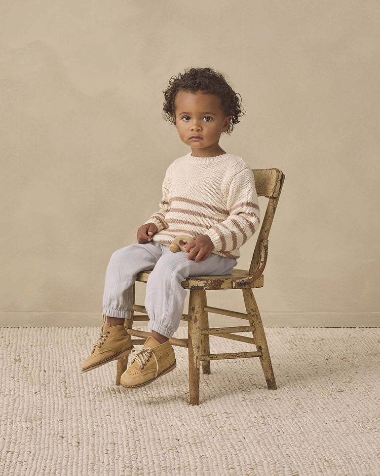A young child with curly hair sits on a worn wooden chair, wearing the quincy mae rex sweater in almond stripe, crafted from soft organic cotton, paired with light pants and tan moccasins, against a neutral background on a textured rug.