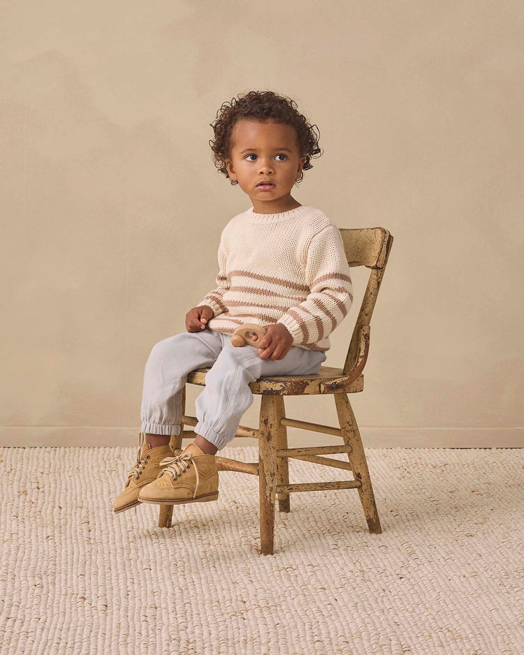 A young child with curly hair sits on a worn wooden chair, wearing the quincy mae rex sweater in almond stripe—an organic cotton style featuring ribbed detailing—paired with light pants and tan boots, holding a wooden toy against a beige background.