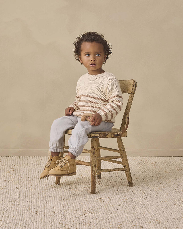 A young child with curly hair sits on a worn wooden chair, wearing the quincy mae rex sweater in almond stripe—an organic cotton style featuring ribbed detailing—paired with light pants and tan boots, holding a wooden toy against a beige background.