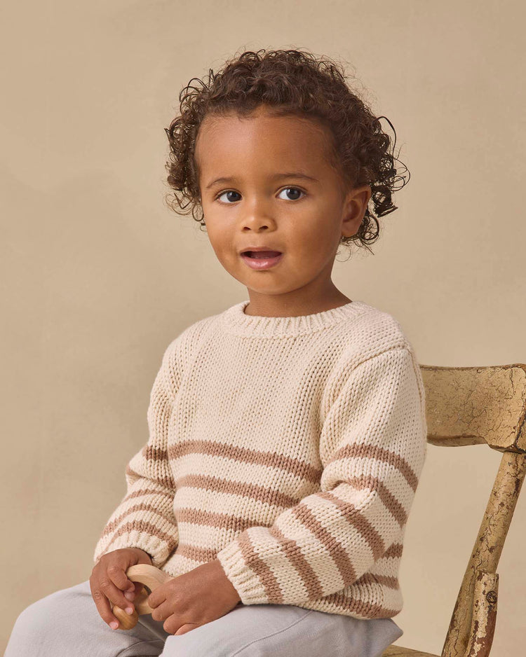 A young child with curly hair sits on a wooden chair, wearing the quincy mae rex sweater in almond stripe and light pants, holding a small object and gazing slightly to the side with a neutral expression.