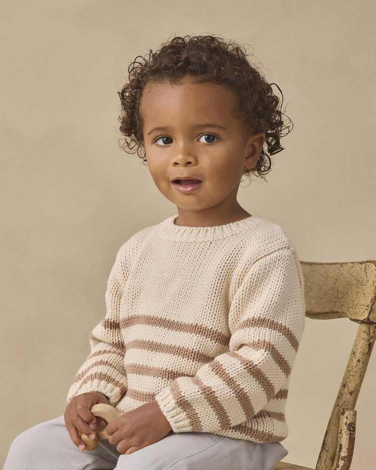 A young child with curly hair sits on a wooden chair, wearing the quincy mae rex sweater in almond stripe and light pants, holding a small object and gazing slightly to the side with a neutral expression.