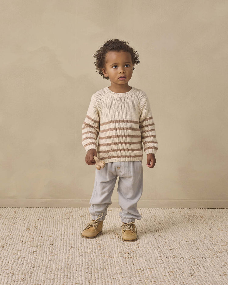 A young child with curly hair stands on a textured rug, wearing the quincy mae rex sweater in almond stripe with ribbed detailing, light pants, and tan shoes, holding a small wooden toy.