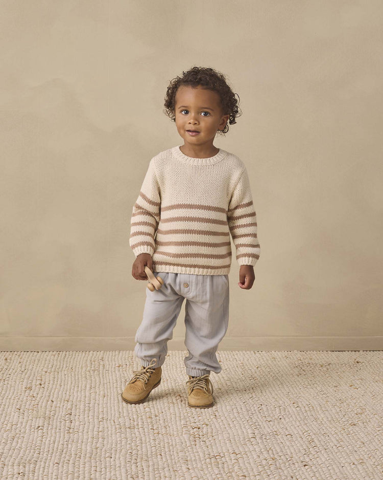 A young child with curly hair stands on a light rug against a beige background, wearing the quincy mae rex sweater in almond stripe crafted from organic cotton, paired with light gray pants and tan shoes, and holding a small wooden toy.