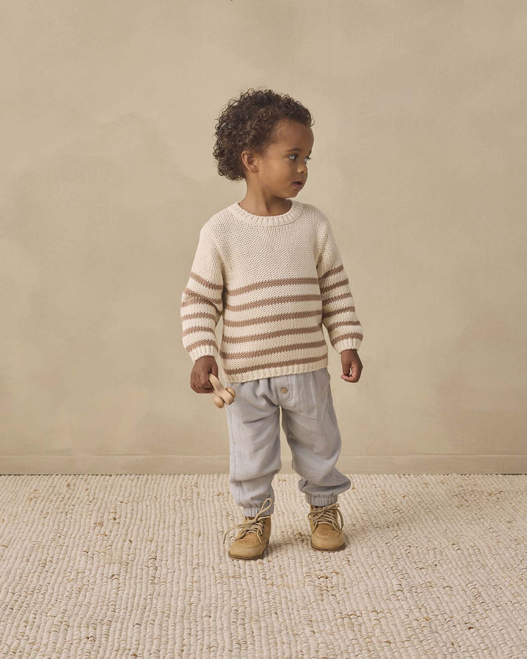 A young child stands on a textured rug, wearing the quincy mae rex sweater in almond stripe, paired with light blue pants and tan boots, holding a small wooden toy and looking to the side against a beige background.