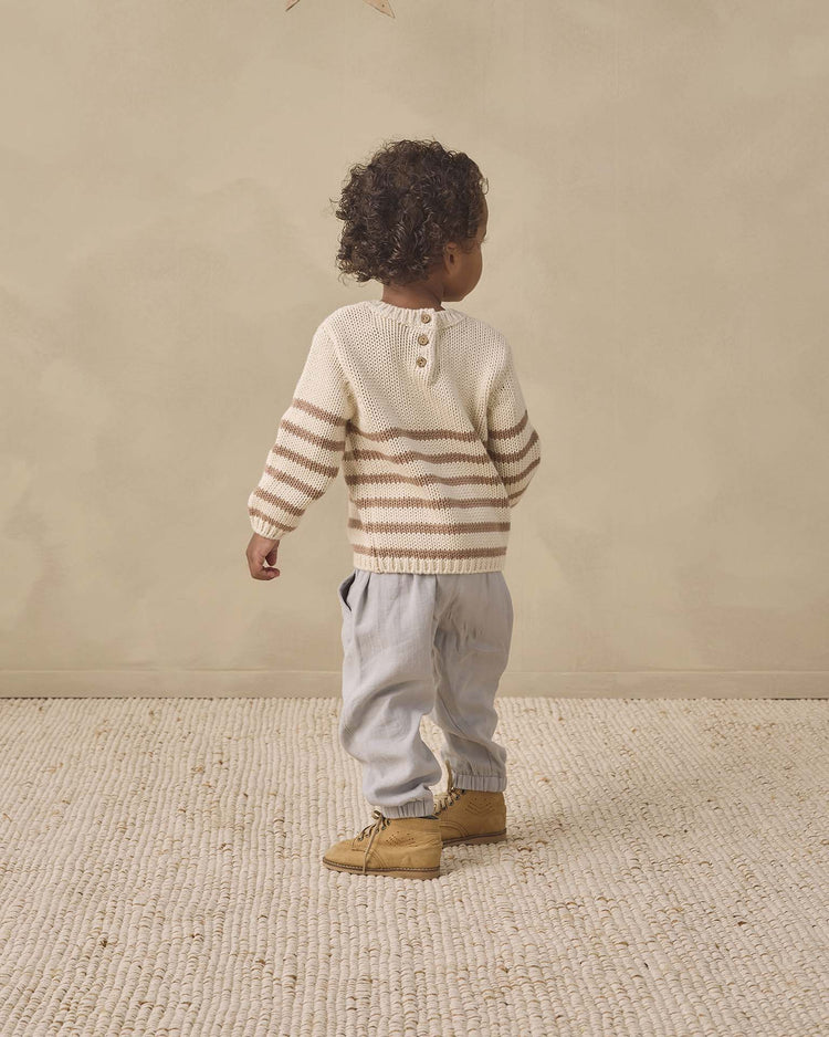 A young child with curly hair stands on a textured rug, facing away. The child wears the quincy mae rex sweater in almond stripe—crafted from organic cotton—paired with light gray pants and tan boots, set against a neutral beige background.