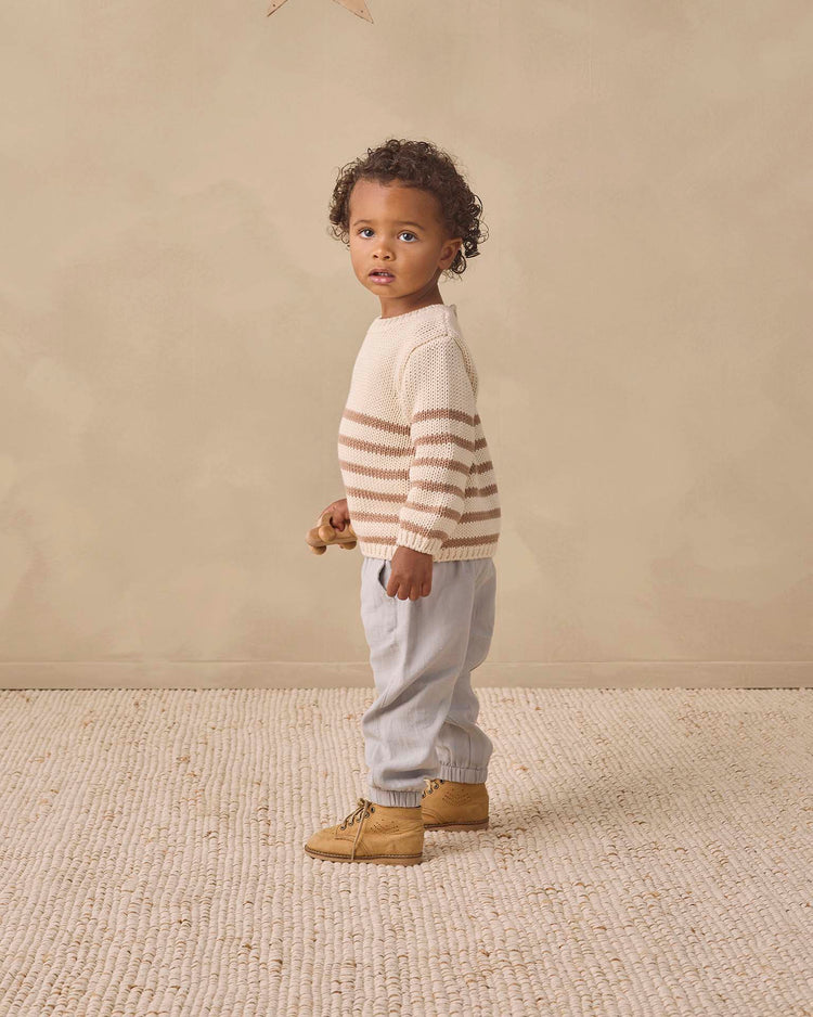 A young child with curly hair stands on a textured carpet, wearing the Quincy Mae Rex Sweater in almond stripe made from soft organic cotton, paired with light blue pants and tan boots, gazing slightly to the side with a neutral expression.