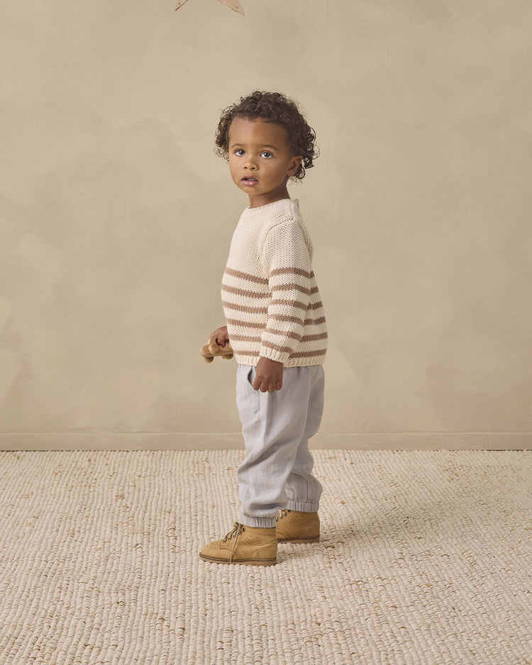 A young child with curly hair stands on a textured carpet, wearing the Quincy Mae Rex Sweater in almond stripe made from soft organic cotton, paired with light blue pants and tan boots, gazing slightly to the side with a neutral expression.