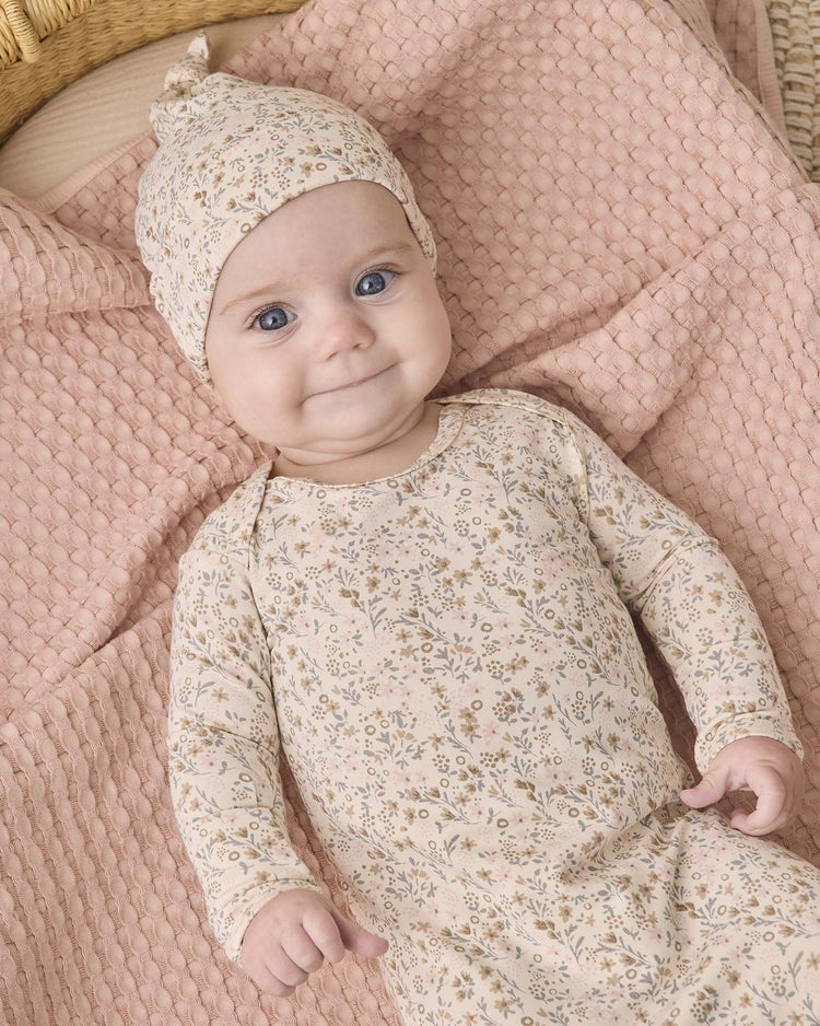 A baby with wide blue eyes and a small smile lies on a textured, light pink blanket, dressed in the quincy mae bamboo baby gown + hat set in bloom, featuring a matching floral-patterned elastic bottom gown and knotted hat.